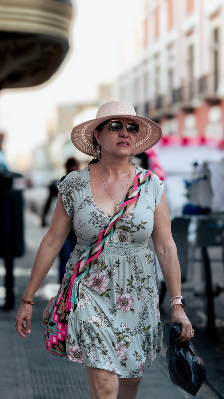 A fashionable woman in a floral dress and hat walking confidently on a sunny city street.